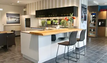 Moderne Ausstellungsküche mit weißer Kochinsel, massiver Holztheke, schwarzen Oberschränken, integrierter Spüle und zwei Barhockern in offenem, stilvoll gestaltetem Showroom.